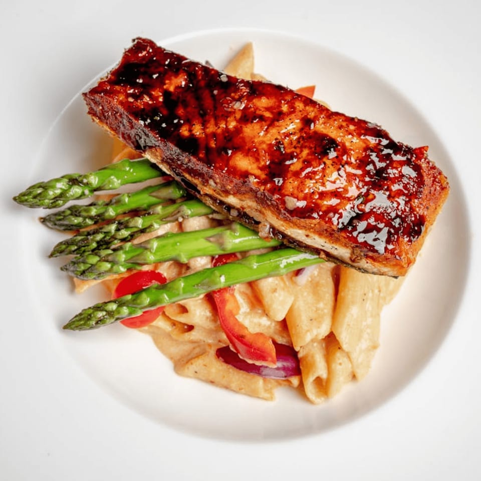Glazed salmon fillet over penne pasta with red peppers and asparagus on a white plate.